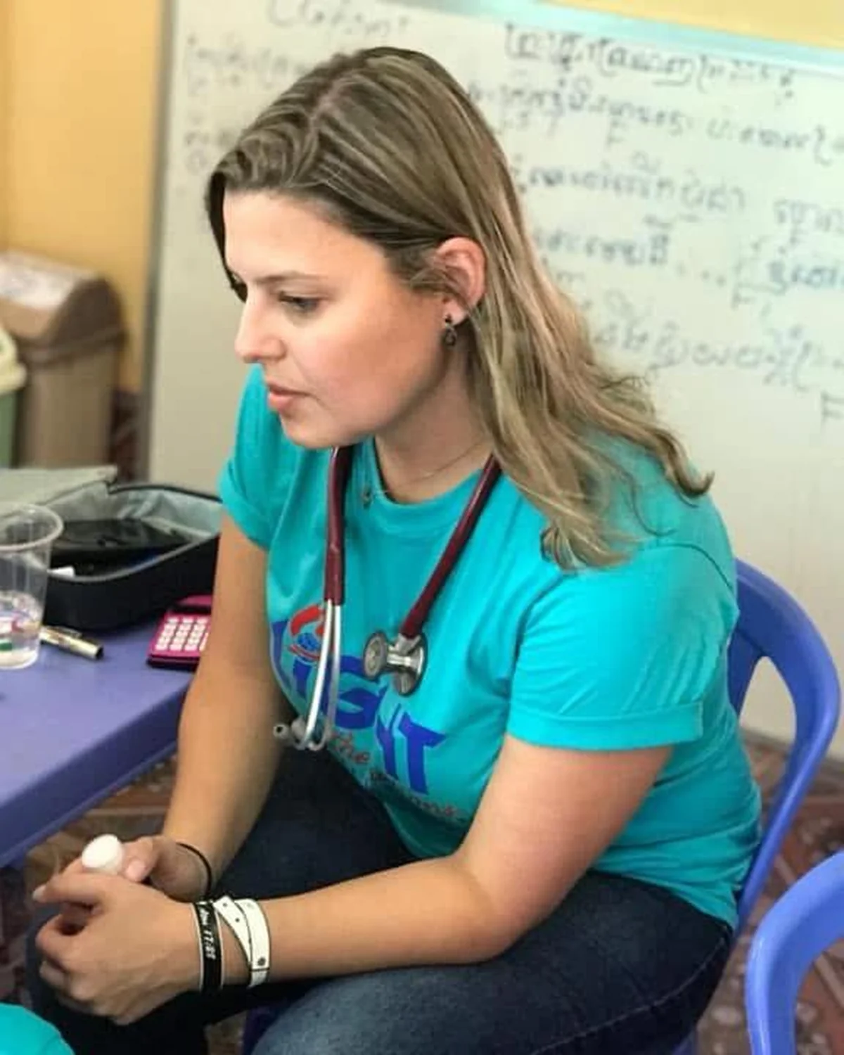 Dra. Tatiana atende uma criança durante uma missão médica, usando estetoscópio e camiseta turquesa, em atenção cuidadosa.