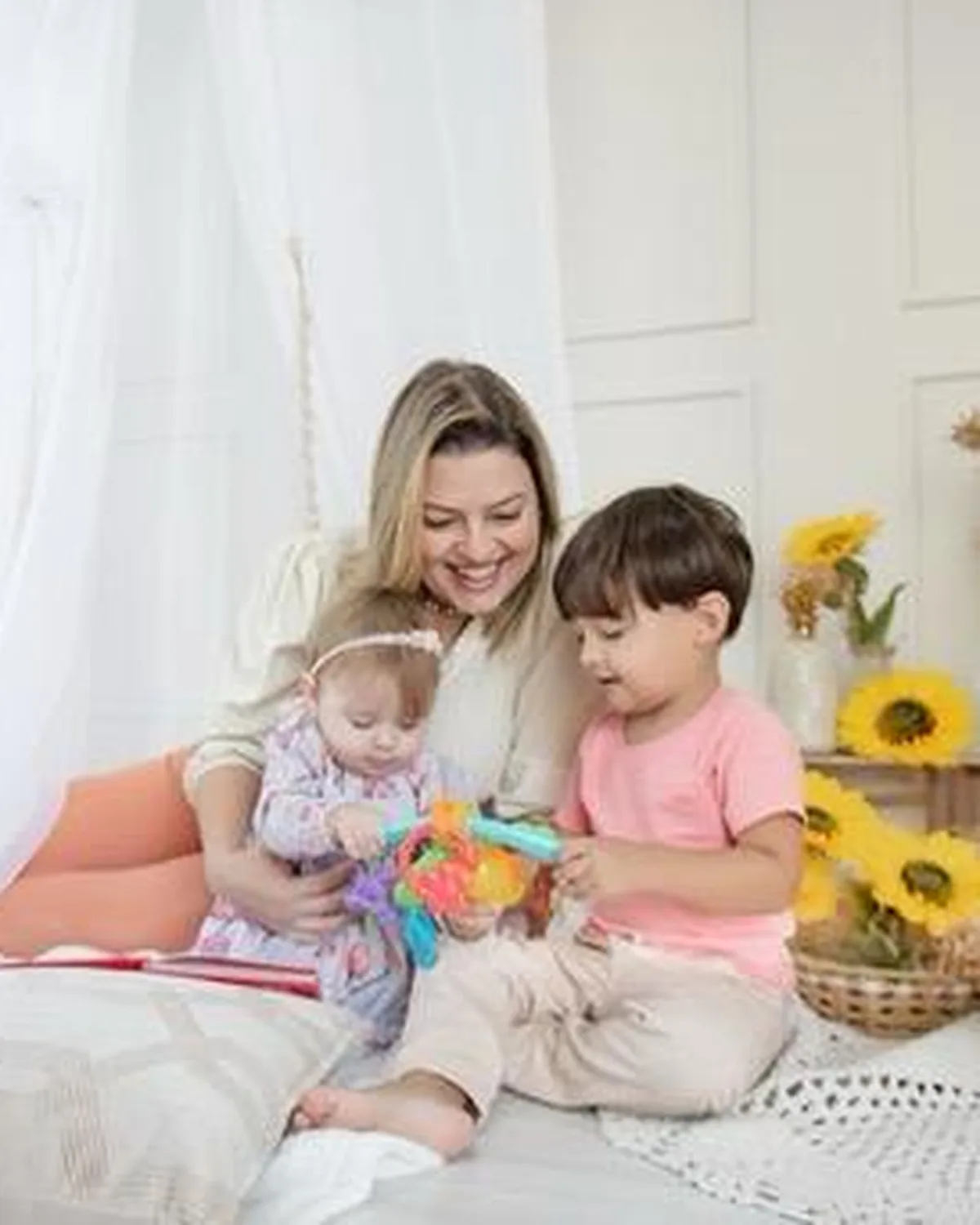 Dra. Tatiana sentada na cama com os dois filhos, lendo e brincando em um quarto claro com girassóis.