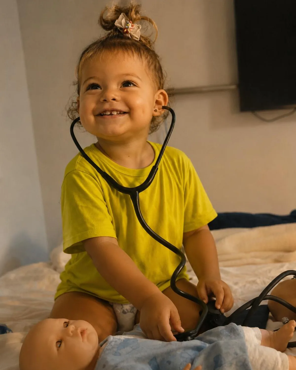 Criança pequena sorrindo com um estetoscópio, brincando de examinar um boneco sobre a cama.