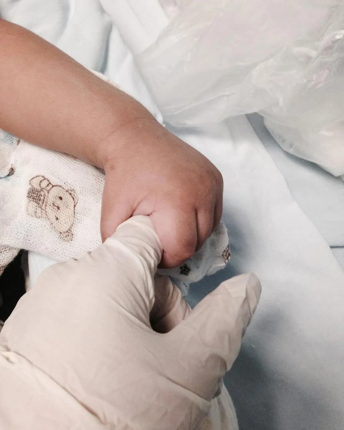 Mão enluvada segurando com cuidado a mãozinha de um bebê em um leito hospitalar.