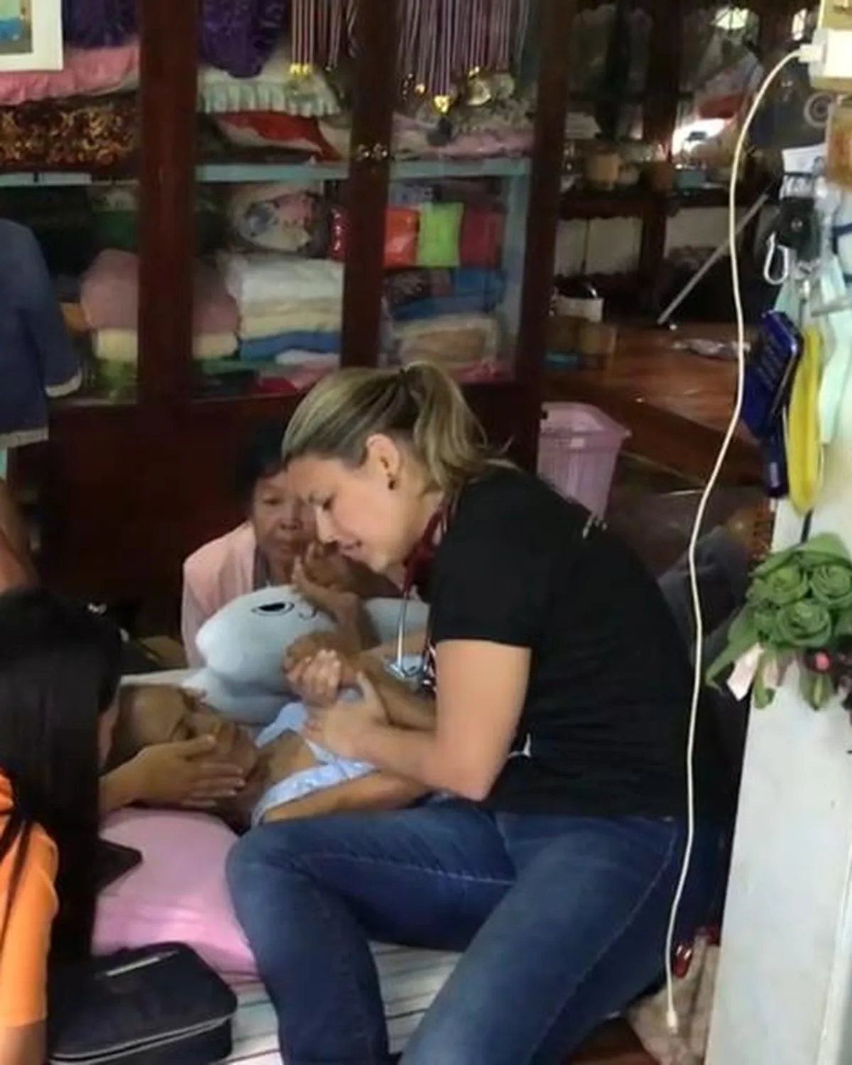 Dra. Tatiana atende um bebê no colo da família dentro de uma casa simples, durante uma missão médica.