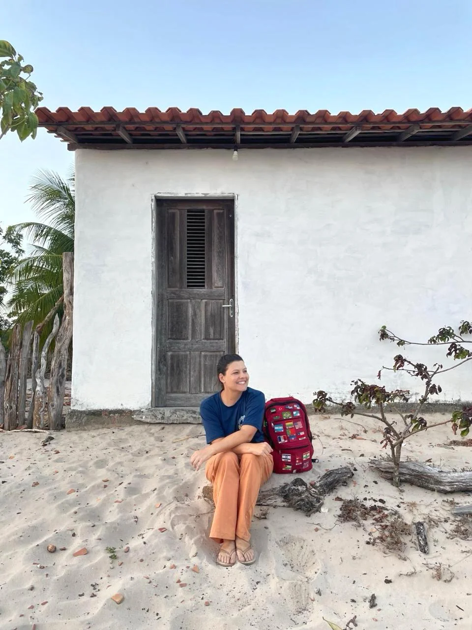 Dra. Tatiana sentada em frente a uma casa simples de areia, com mochila vermelha ao lado, contemplando o horizonte durante uma missão